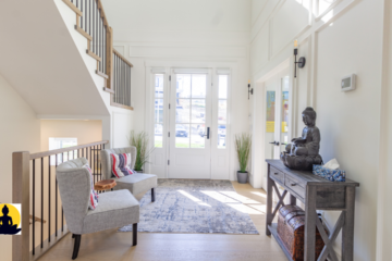 The image shows a modern, serene entryway of a home, filled with natural light coming through a set of large French doors. The room is decorated in neutral tones, with white walls and light wood flooring, creating a peaceful and inviting atmosphere. A wooden console table stands against the wall on the right, and on top of it sits a dark-colored Buddha statue in a meditative pose. The statue adds a calming spiritual presence to the space. Two grey accent chairs with red and white pillows are arranged beside a small wooden side table under the staircase on the left. A soft, patterned rug lies on the floor, tying the space together. There are a few potted plants placed strategically, bringing a touch of nature indoors. A small Buddha icon appears in the bottom-left corner, possibly as a logo or branding element. The overall ambiance is tranquil and harmonious.